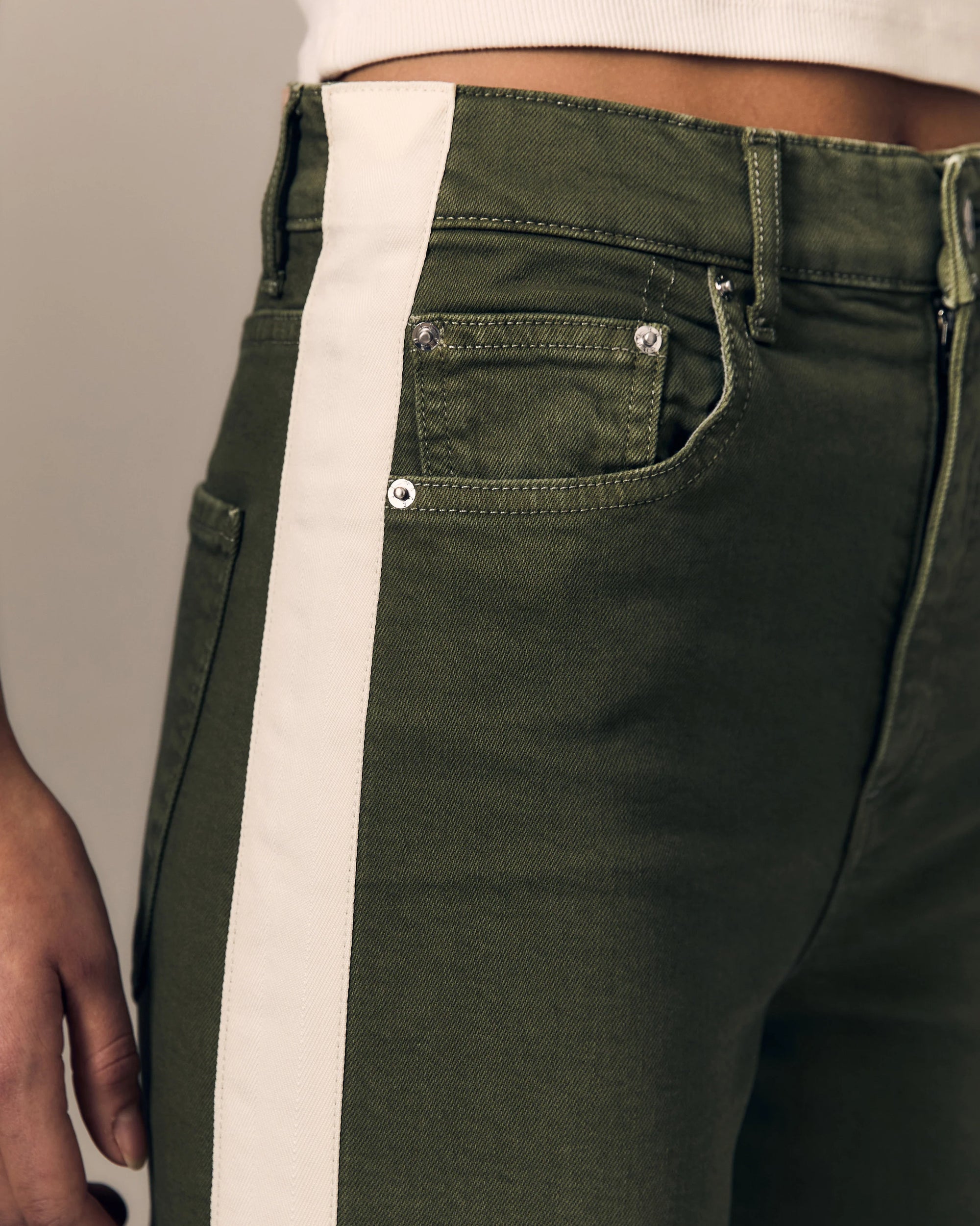 Close-up of olive green denim jeans with a wide off-white vertical stripe running down the side, worn by a model.