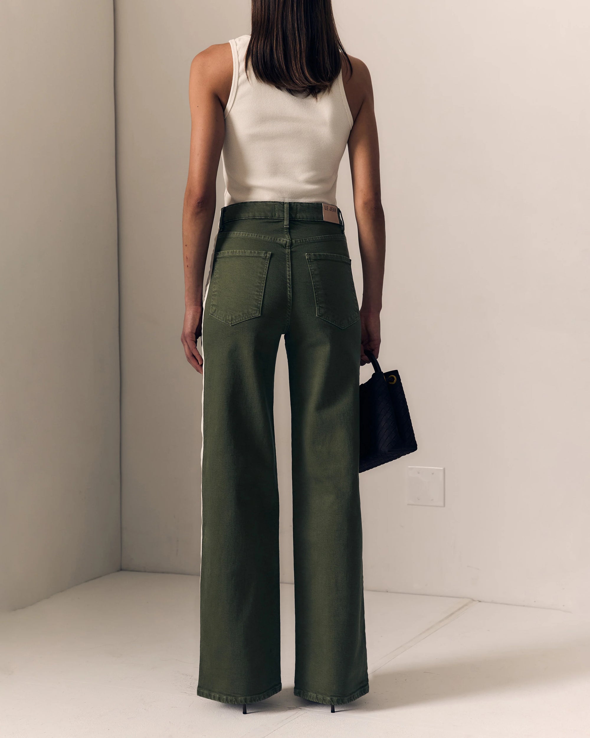 A model shown from the back wearing olive green wide-leg pants with a white ribbed tank top and holding a black handbag, set against a minimalist beige studio background.