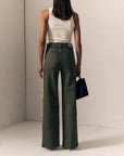 A model shown from the back wearing olive green wide-leg pants with a white ribbed tank top and holding a black handbag, set against a minimalist beige studio background.