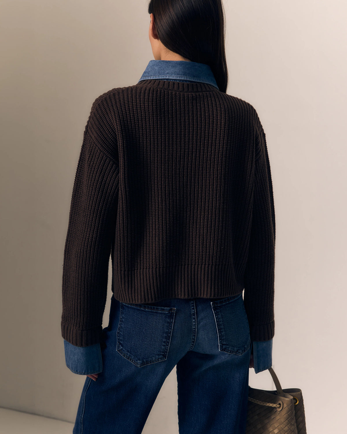 model wearing the harvey sweater that is a brown ribbed sweater with denim collar and cuff trim with jeans on a cream background, back view