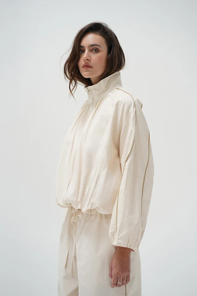 A woman wearing a cream-colored lightweight jacket with a high collar and matching drawstring pants, photographed against a plain light gray studio background.