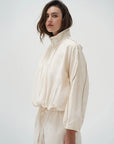 A woman wearing a cream-colored lightweight jacket with a high collar and matching drawstring pants, photographed against a plain light gray studio background.