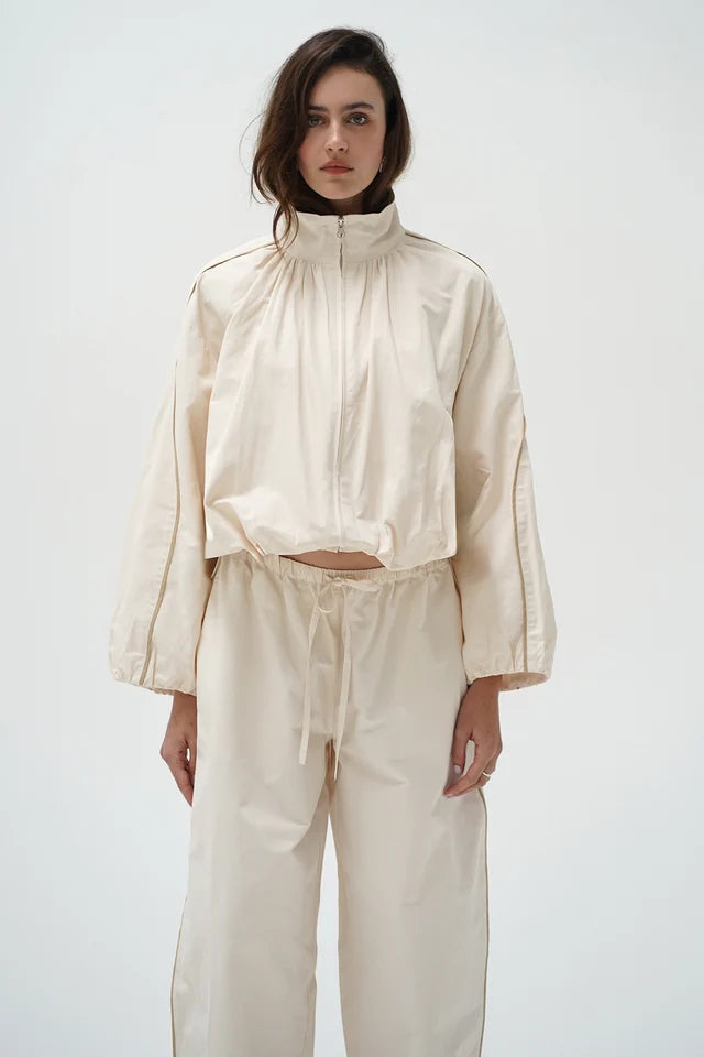 A model wearing a matching cream-colored two-piece outfit consisting of a cropped long-sleeve zip-up jacket with a high collar and matching wide-leg drawstring pants, set against a plain light gray background.