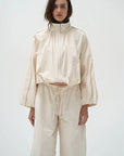A model wearing a matching cream-colored two-piece outfit consisting of a cropped long-sleeve zip-up jacket with a high collar and matching wide-leg drawstring pants, set against a plain light gray background.