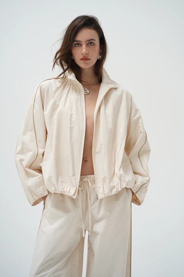 A model wearing a light cream/off-white zip-up jacket and matching wide-leg pants against a plain light gray studio background.