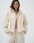 A model wearing a light cream/off-white zip-up jacket and matching wide-leg pants against a plain light gray studio background.