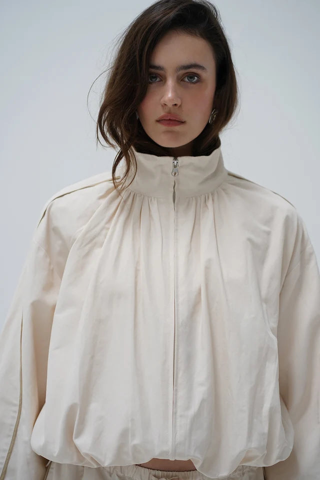 A model wearing a cream-colored, high-neck zip-up jacket with dramatic gathered fabric and wide sleeves, set against a plain light gray background.