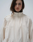 A model wearing a cream-colored, high-neck zip-up jacket with dramatic gathered fabric and wide sleeves, set against a plain light gray background.