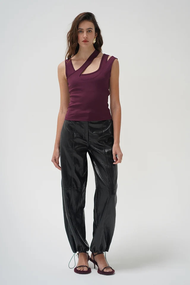 A model stands against a light gray studio background wearing a sleeveless deep purple top with a crossover neckline and black shiny cropped pants with drawstring ankle ties.