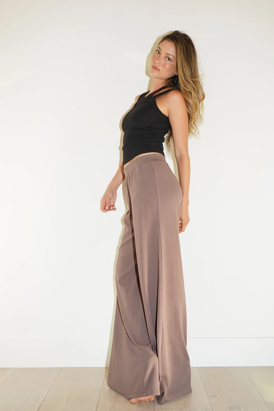 A woman modeling a black halter top and high-waisted, wide-leg taupe maxi pants against a plain white wall and light wood floor.