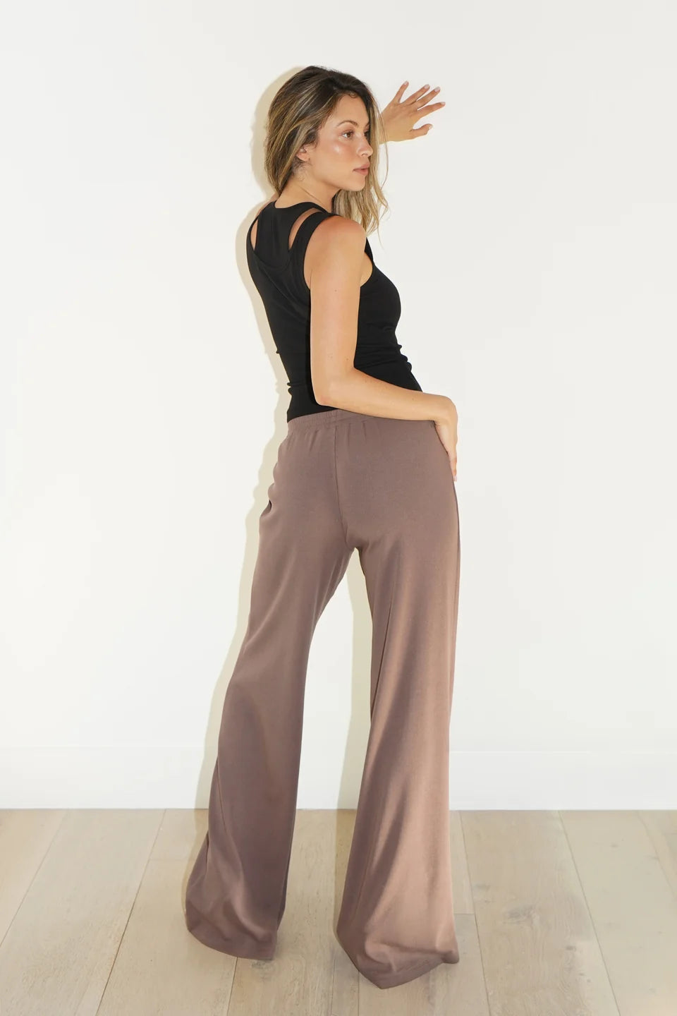 A woman wearing high-waisted, wide-leg taupe pants and a black sleeveless top, standing in profile against a white wall with light wood flooring.
