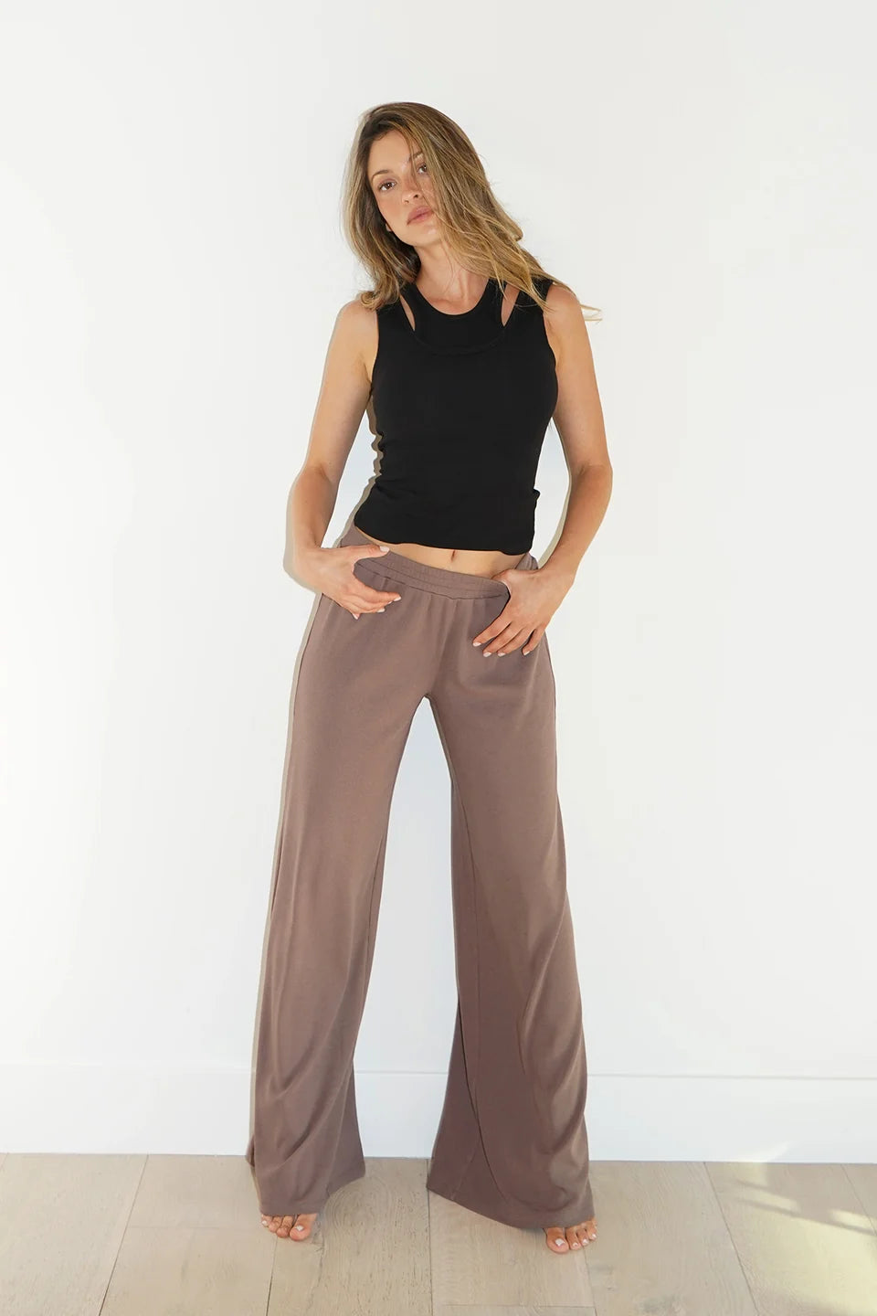 A woman modeling a pair of wide-leg taupe pants with a black sleeveless crop top, standing against a white wall on a light wood floor.