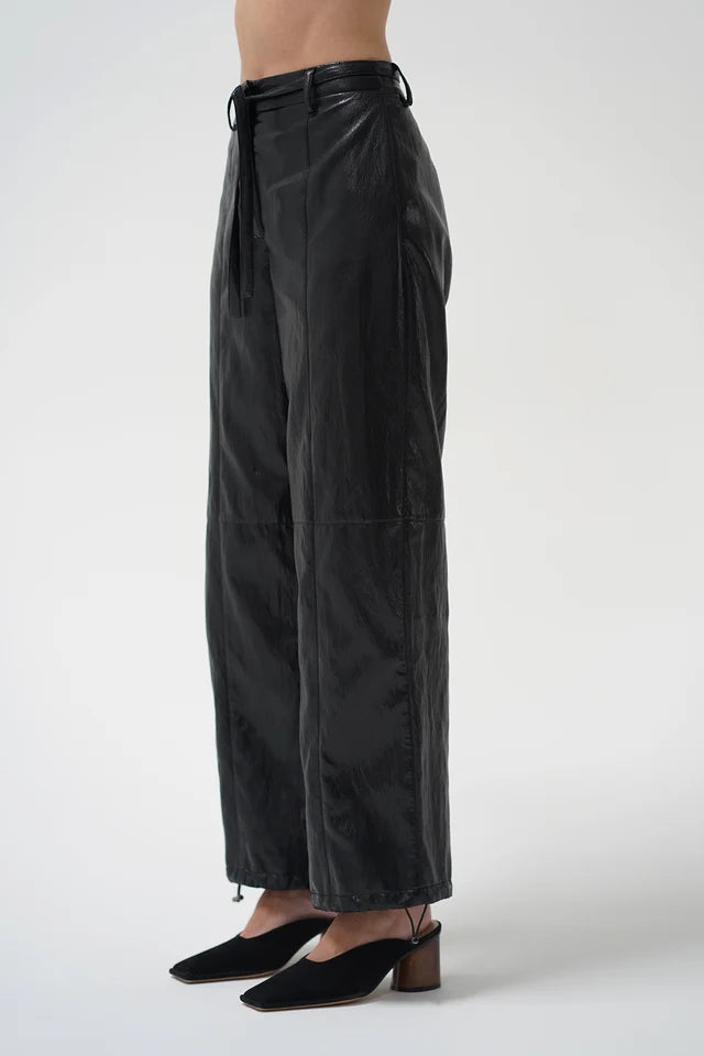 A model is shown from the waist down wearing black wide-leg pants with a drawstring waist and black pointed-toe mule heels, set against a plain light gray studio background.