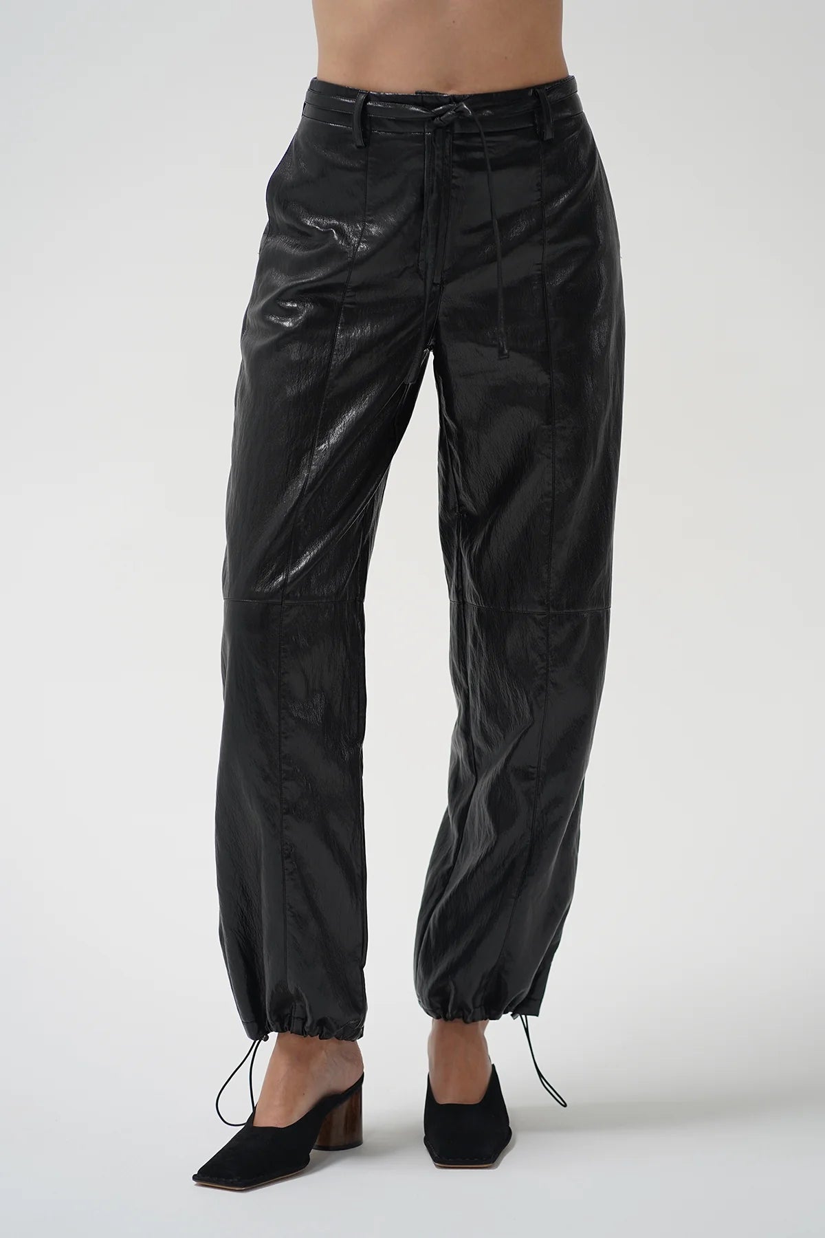 Black glossy cropped pants with a drawstring waist and ankle ties, shown on a model wearing black pointed mule heels against a light gray studio background.