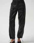 Black glossy cropped pants with a drawstring waist and ankle ties, shown on a model wearing black pointed mule heels against a light gray studio background.