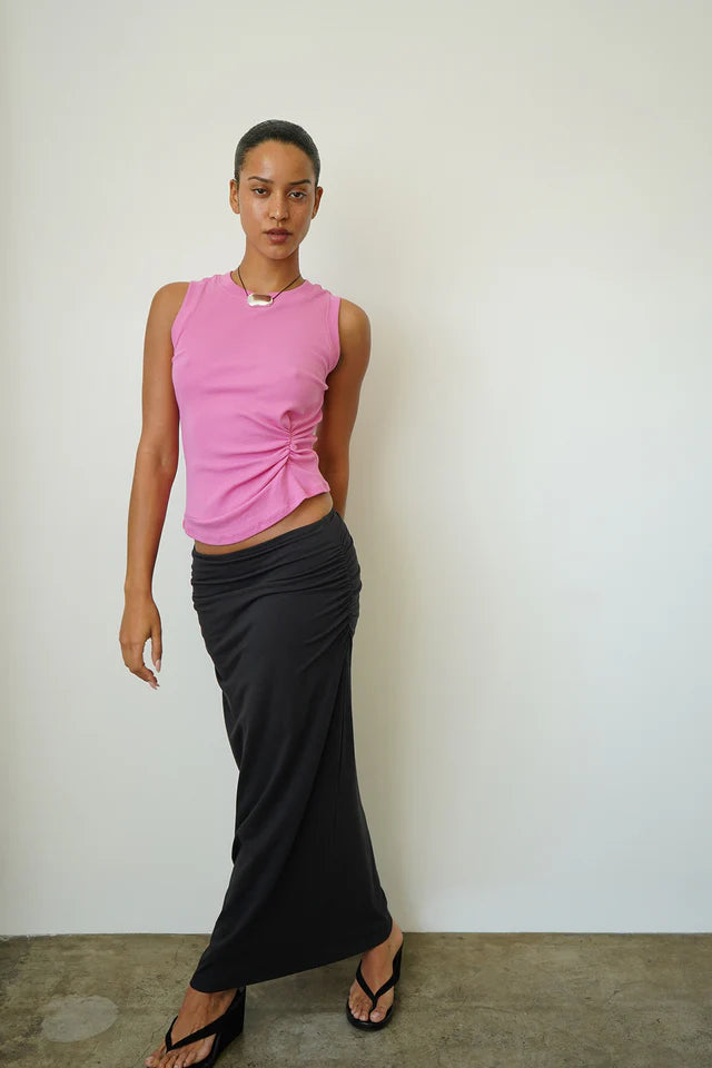 Woman wearing a pink sleeveless top and black skirt against a white wall.