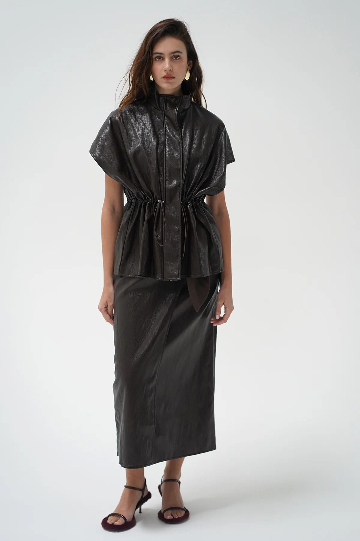 A model wearing a dark brown leather top and a matching skirt, paired with dark heeled sandals, against a plain white studio background.