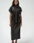 A model wearing a dark brown leather top and a matching skirt, paired with dark heeled sandals, against a plain white studio background.