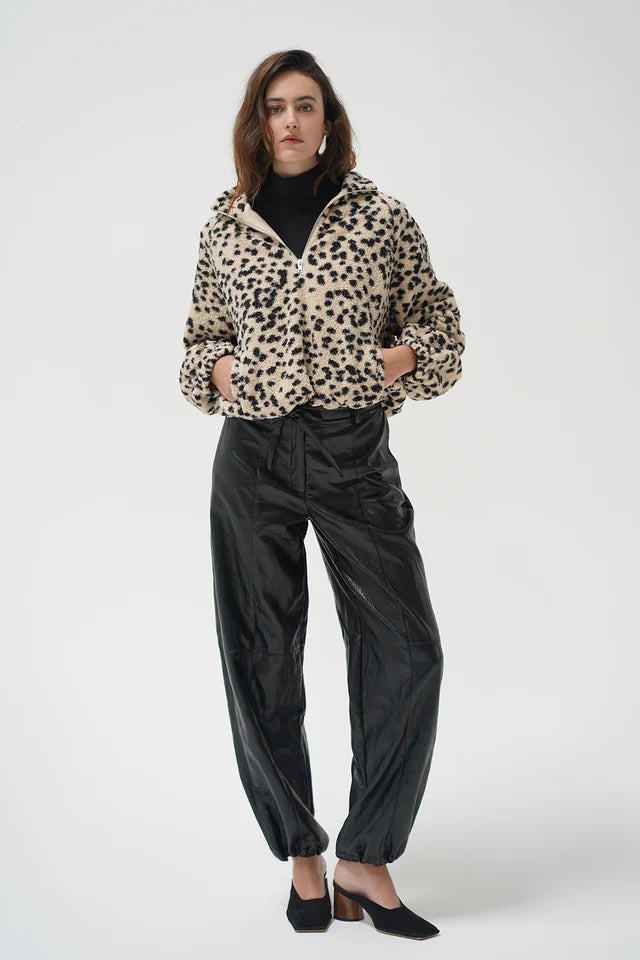 A model stands against a light gray studio background wearing a beige and black leopard-print cropped jacket over a black turtleneck, paired with black satin trousers and black pointed-toe heels.