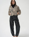 A model stands against a light gray studio background wearing a beige and black leopard-print cropped jacket over a black turtleneck, paired with black satin trousers and black pointed-toe heels.