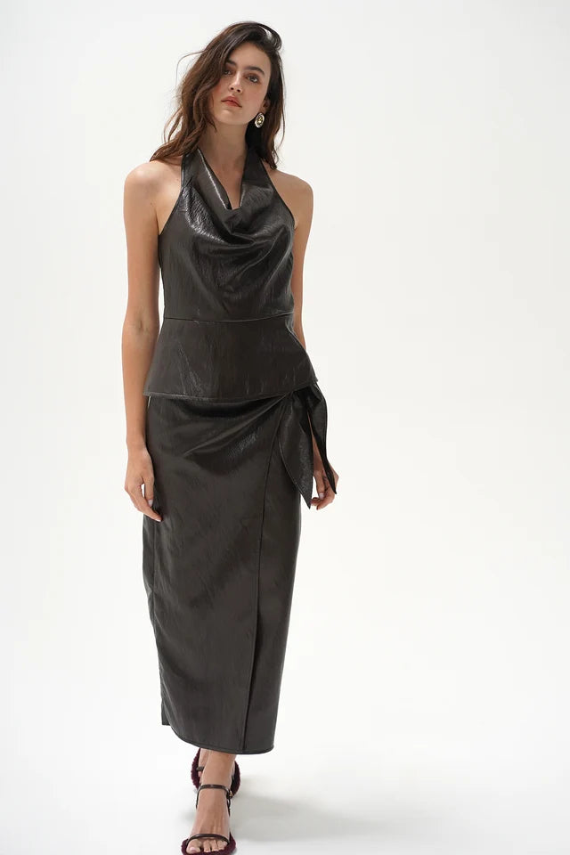 A model wearing a dark brown leather halter-neck wrap top with a draped cowl neckline and a matching skirt, paired with dark heeled sandals, against a plain white studio background.