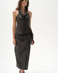 A model wearing a dark brown leather halter-neck wrap top with a draped cowl neckline and a matching skirt, paired with dark heeled sandals, against a plain white studio background.