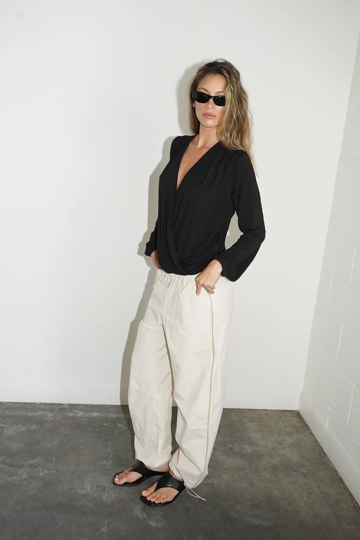 A woman modeling a black long-sleeve wrap-style top and cream wide-leg pants with a thin gold zipper detail, standing in a minimalist room with a white wall and gray concrete floor.