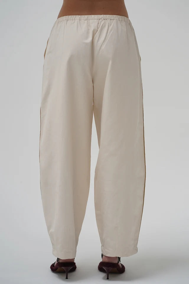 A pair of loose-fitting, cream-colored wide-leg pants with side pockets and a drawstring waist, shown from the back on a model wearing dark brown heels against a light gray background.
