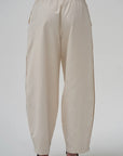 A pair of loose-fitting, cream-colored wide-leg pants with side pockets and a drawstring waist, shown from the back on a model wearing dark brown heels against a light gray background.