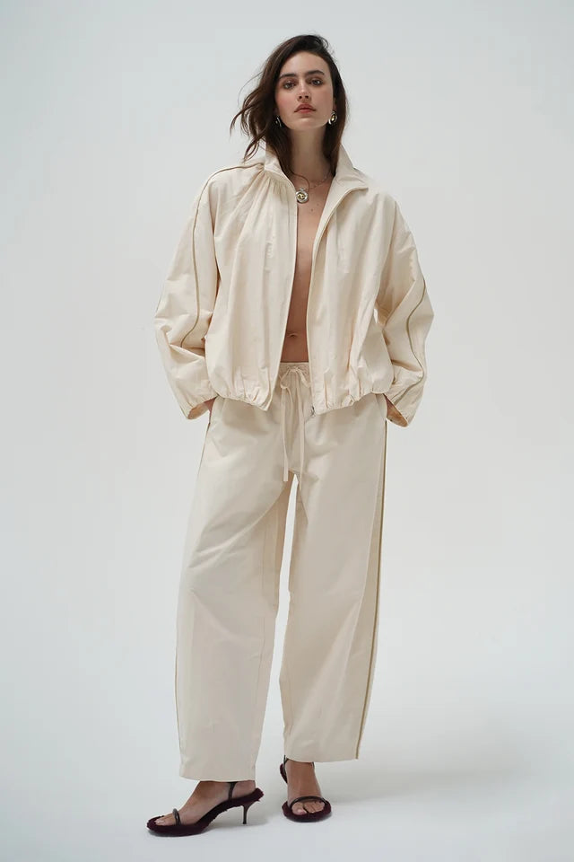 A model wearing a matching cream-colored two-piece set consisting of a lightweight zip-up jacket and wide-leg trousers, styled with dark brown heeled sandals against a plain light gray background.