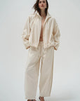 A model wearing a matching cream-colored two-piece set consisting of a lightweight zip-up jacket and wide-leg trousers, styled with dark brown heeled sandals against a plain light gray background.