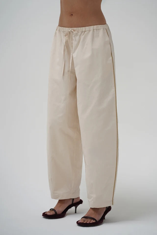 A person wearing loose, wide-leg beige drawstring pants with dark heeled sandals, shown from the waist down against a plain light gray studio background.