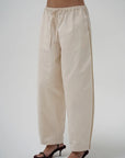A person wearing loose, wide-leg beige drawstring pants with dark heeled sandals, shown from the waist down against a plain light gray studio background.