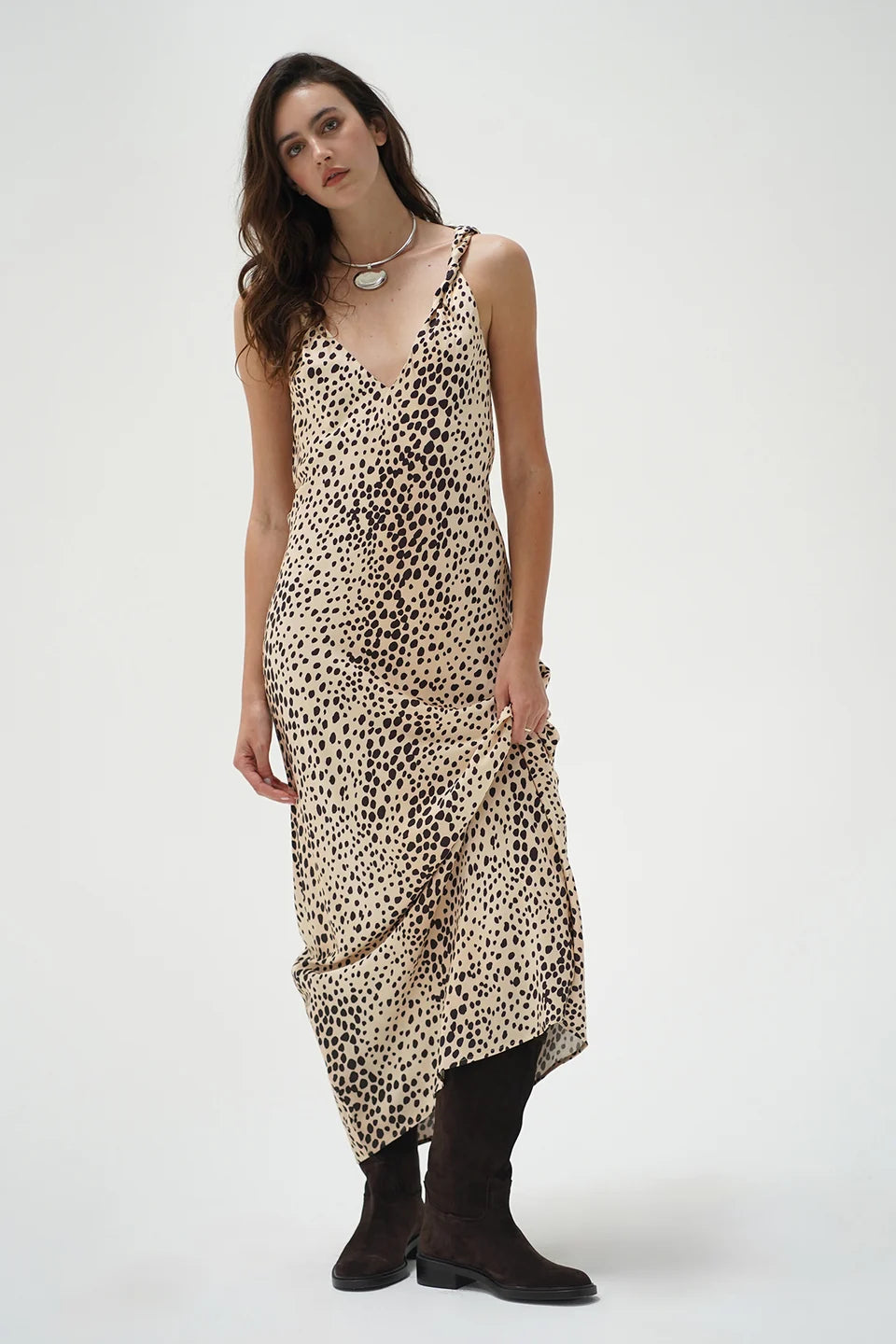 A woman modeling a sleeveless beige slip dress with a black leopard print pattern, paired with black boots, against a plain light gray studio background.