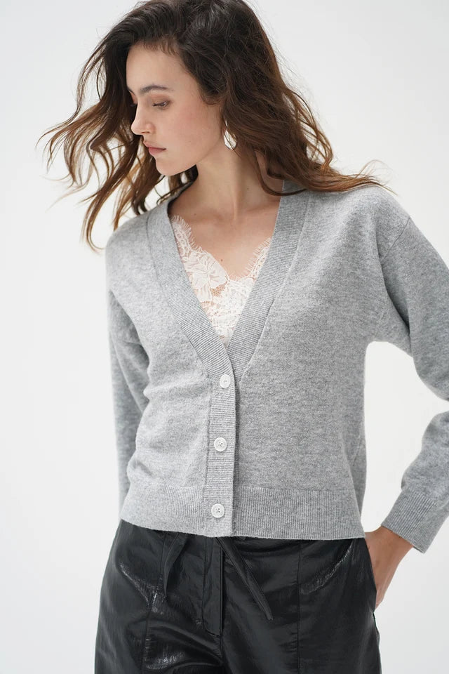 A woman wearing a light gray knit cardigan with white buttons over a white lace top and black pants, photographed against a clean white studio background.