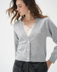 A woman wearing a light gray knit cardigan with white buttons over a white lace top and black pants, photographed against a clean white studio background.