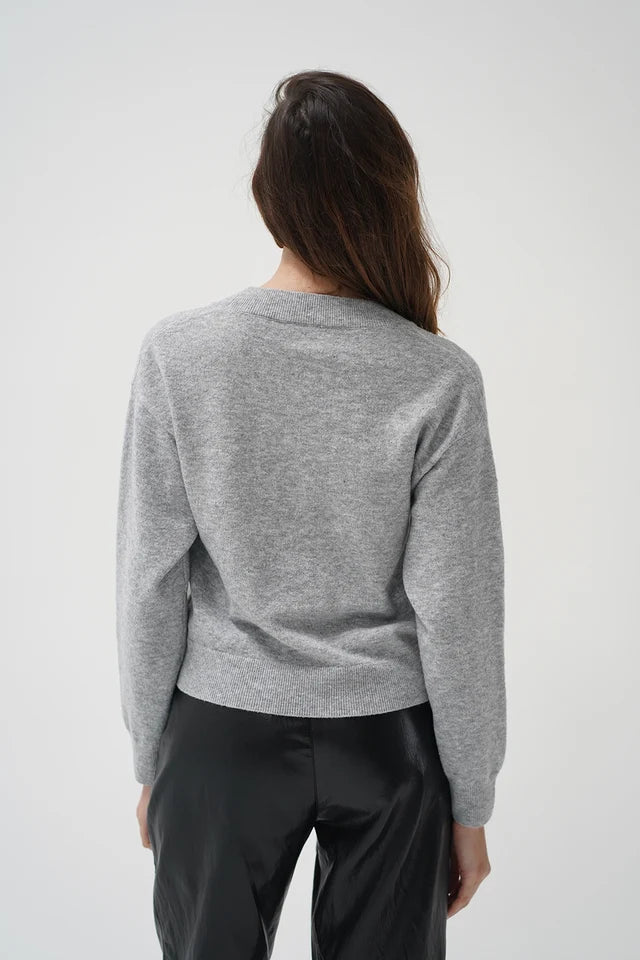 A woman viewed from behind wearing a light gray heathered long-sleeve knit top and black pants, standing against a plain light gray studio background.
