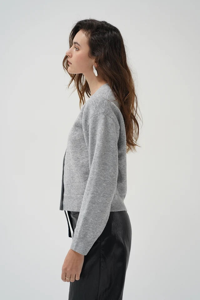 A woman in side profile wearing a light gray cropped knit cardigan and black leather pants against a plain light gray studio background.