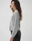 A woman in side profile wearing a light gray cropped knit cardigan and black leather pants against a plain light gray studio background.