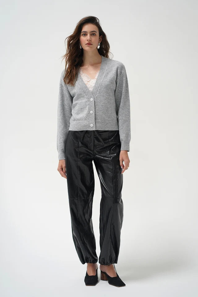 A woman modeling a light gray button-up cardigan, black shiny cropped pants, and black pointed shoes against a plain light gray studio background.
