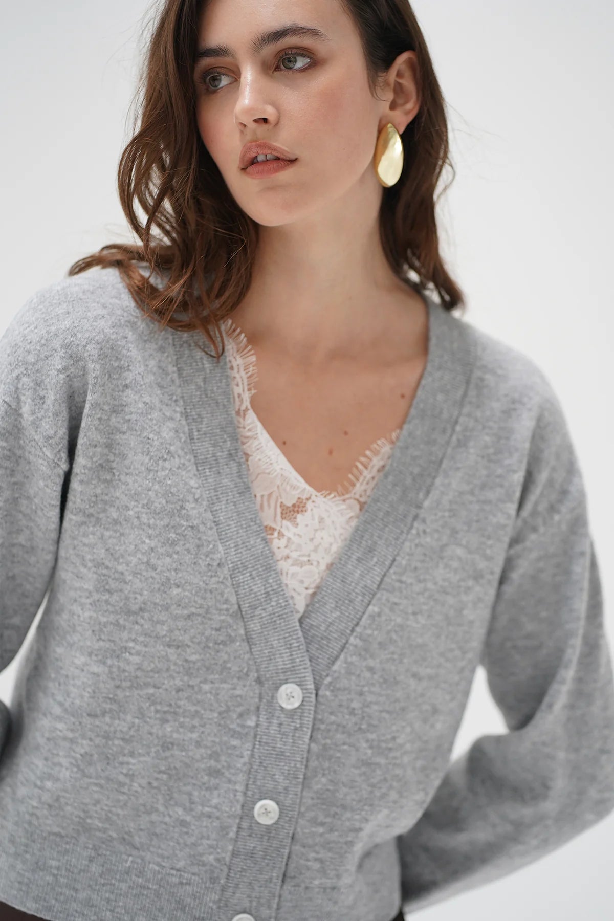 A woman wearing a light gray knit cardigan over a white lace top, photographed against a clean light gray studio background.