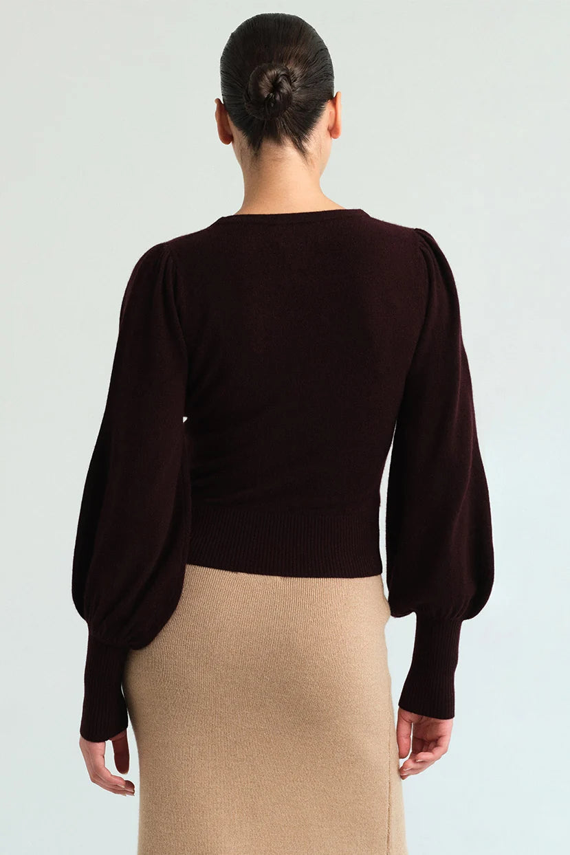 A woman shown from the back wearing a dark brown long-sleeve knit top with balloon sleeves and a beige skirt, against a light gray studio background.