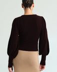 A woman shown from the back wearing a dark brown long-sleeve knit top with balloon sleeves and a beige skirt, against a light gray studio background.