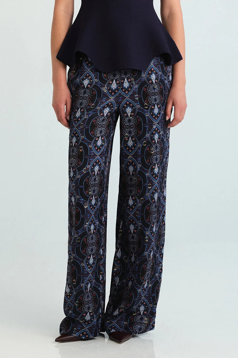 A model is shown from the waist down wearing wide-leg trousers with an intricate blue, black, and white baroque-style pattern, paired with a dark navy peplum top and dark brown pointed shoes.