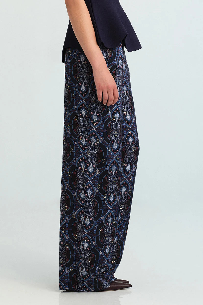 A model is shown from the waist down wearing wide-leg trousers with a detailed blue, black, and white architectural pattern, paired with a dark navy top and brown shoes, against a light gray studio background.