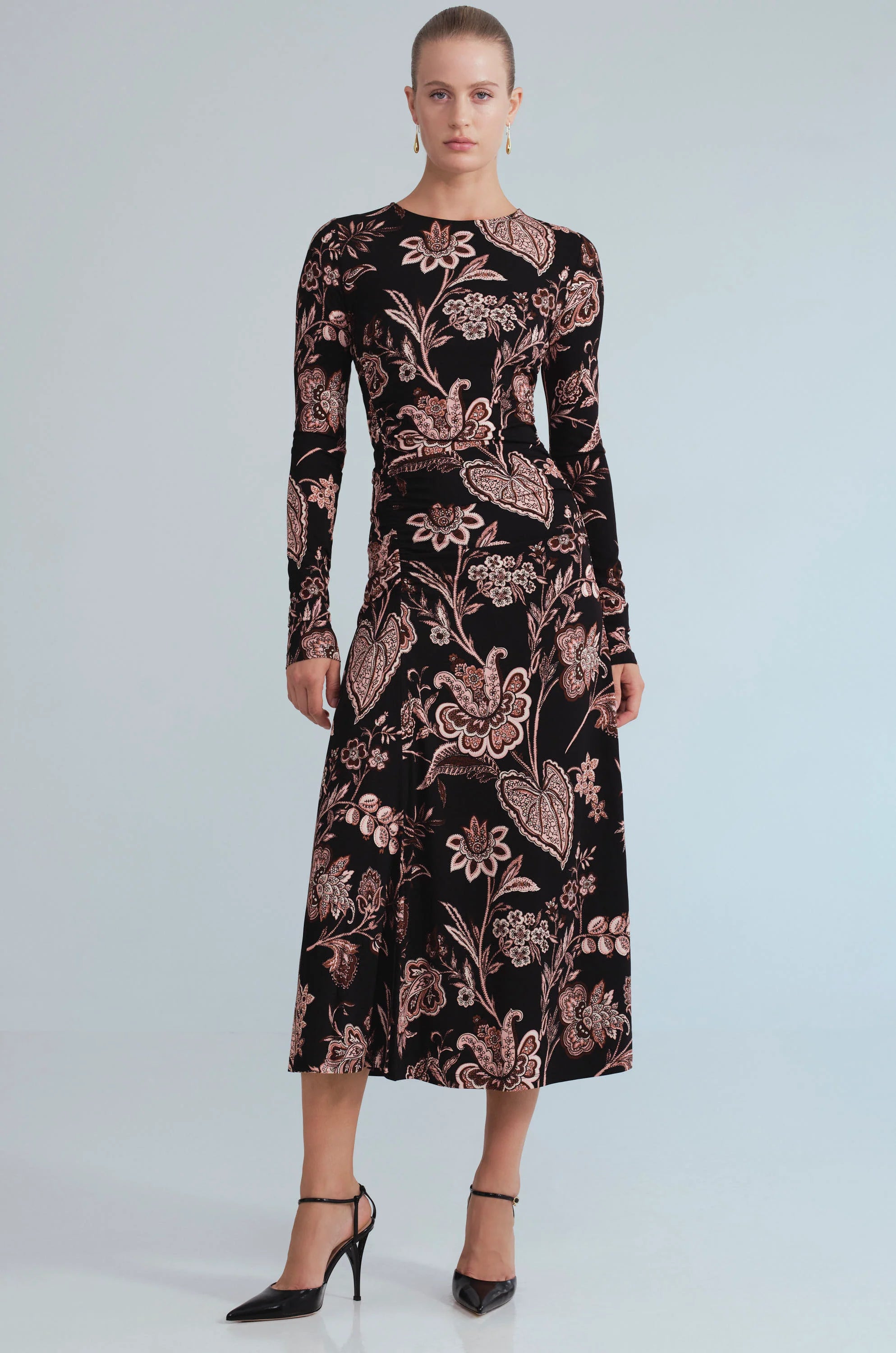 A woman modeling a long-sleeve midi dress with a black base and a dusty rose floral and paisley pattern, paired with black pointed-toe heels against a light gray studio background.