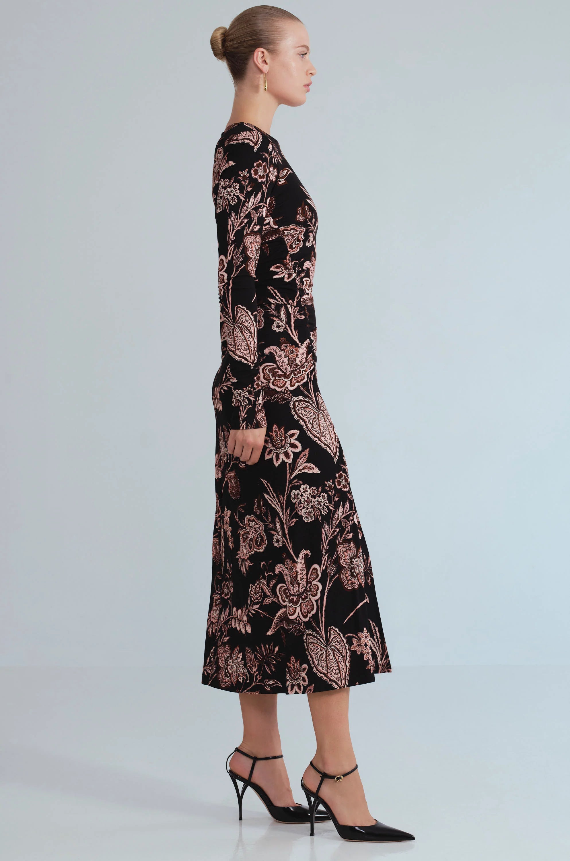 A woman in profile wearing a long-sleeve midi dress with a black base and a dusty rose floral pattern, paired with black high heels, against a light gray studio background.