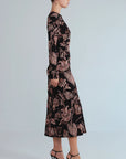 A woman in profile wearing a long-sleeve midi dress with a black base and a dusty rose floral pattern, paired with black high heels, against a light gray studio background.