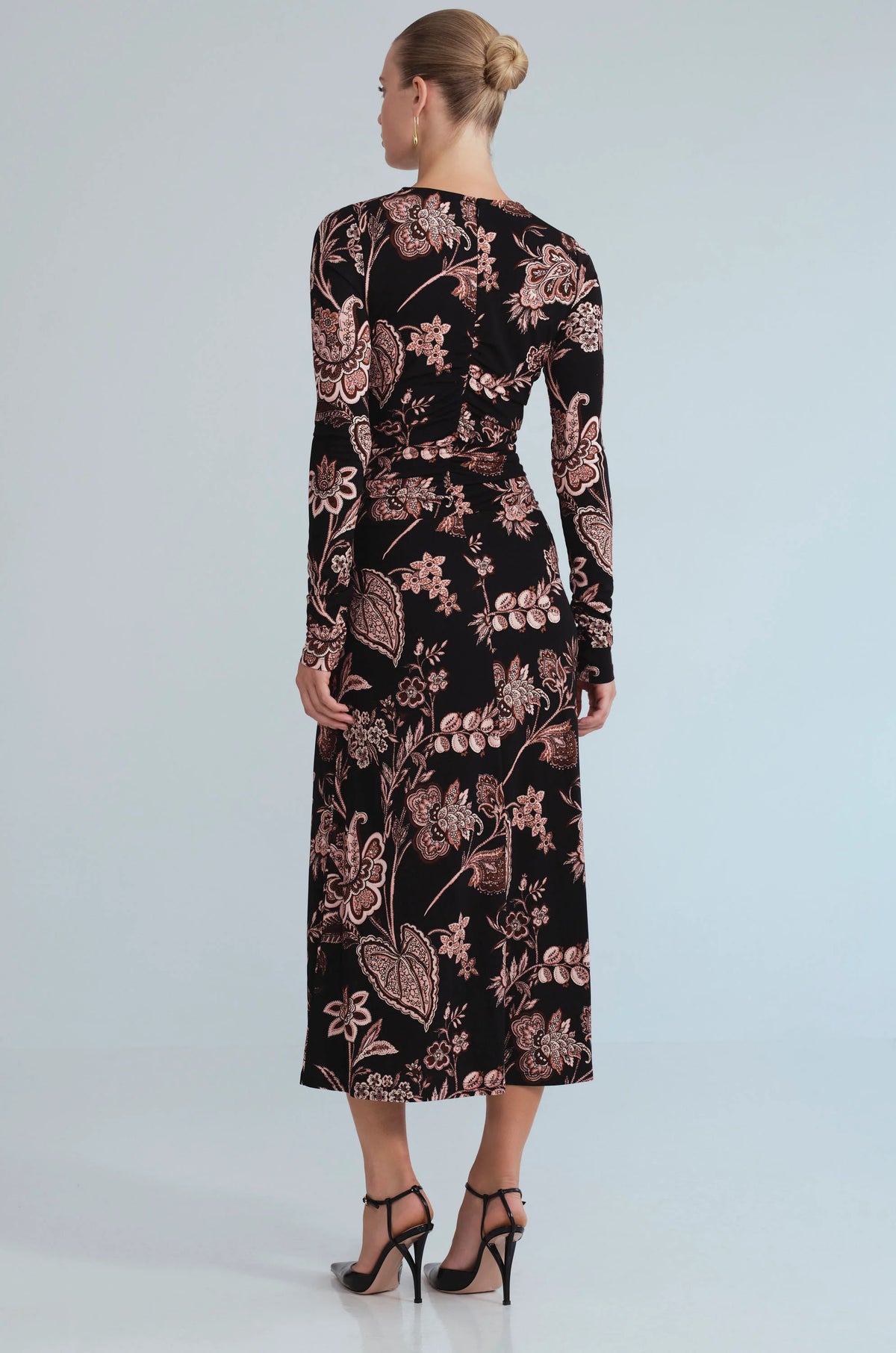 A woman viewed from the back wearing a long-sleeve midi dress with a black base and a dusty rose floral pattern, paired with black high-heeled sandals against a light gray studio background.
