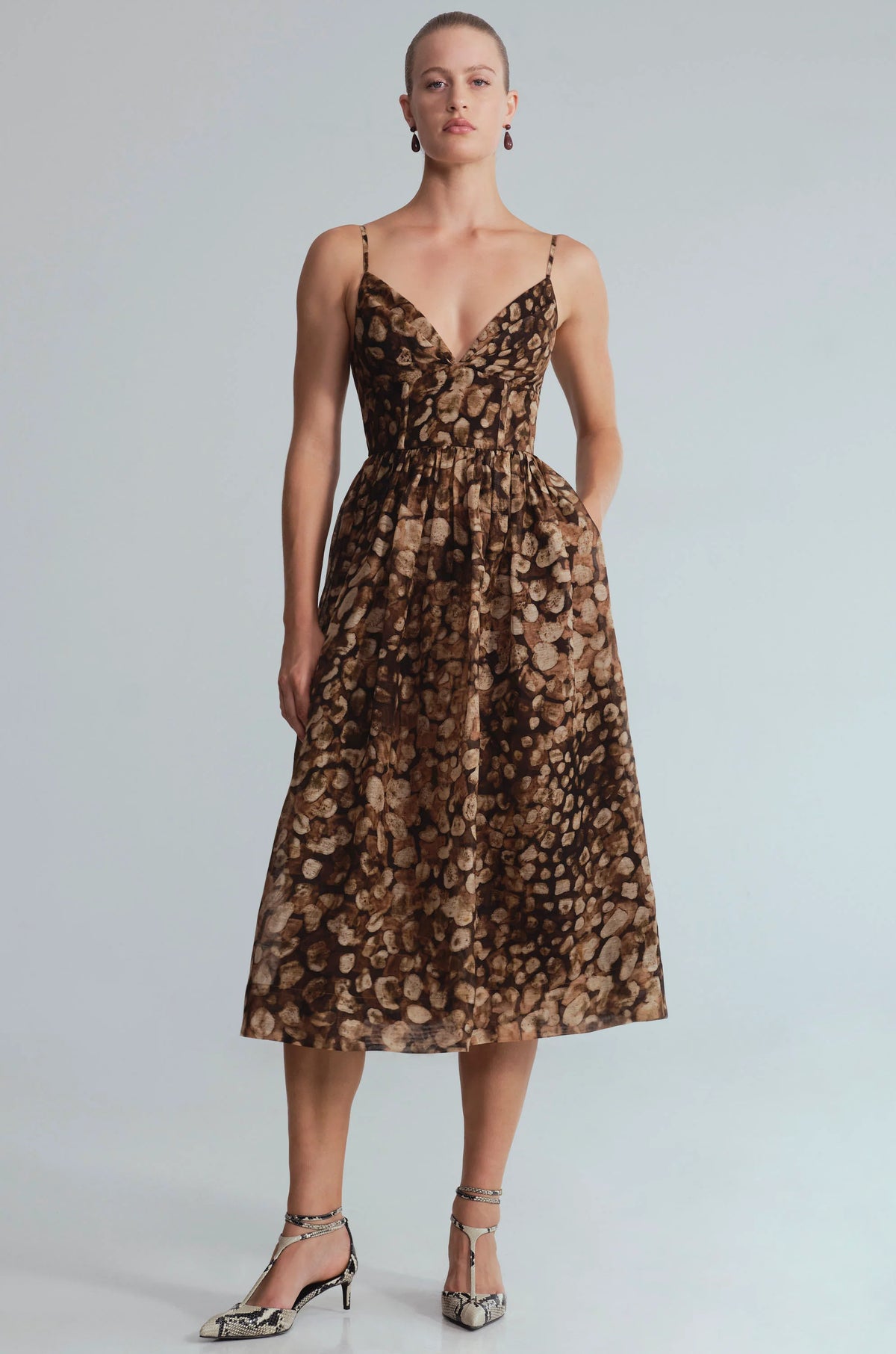 A woman models a brown and beige animal-print midi dress with spaghetti straps and a V-neckline, paired with metallic silver high-heeled sandals, against a light gray studio background.